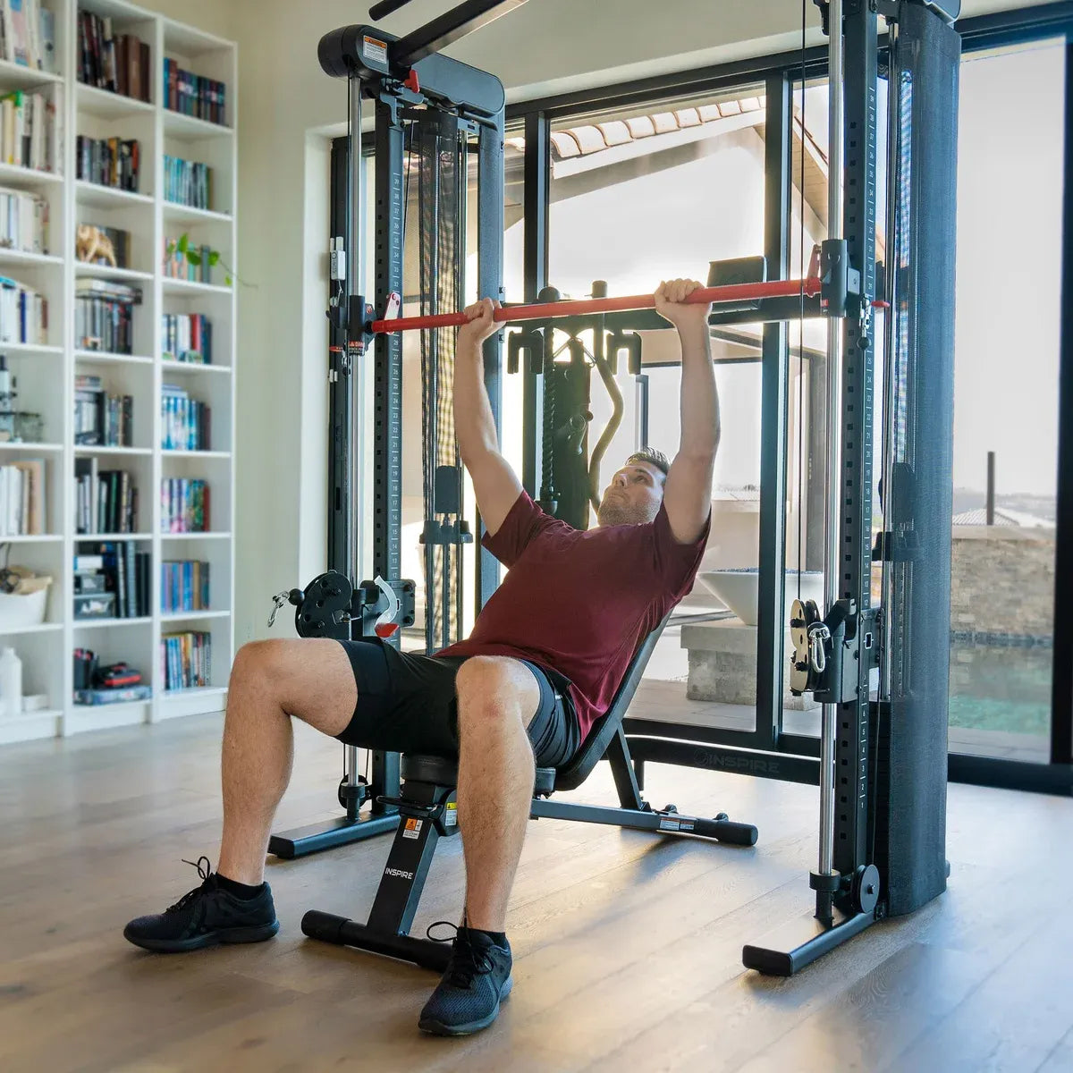 Person exercising on a weight machine in a home setting with bookshelves and a pool in the background.
