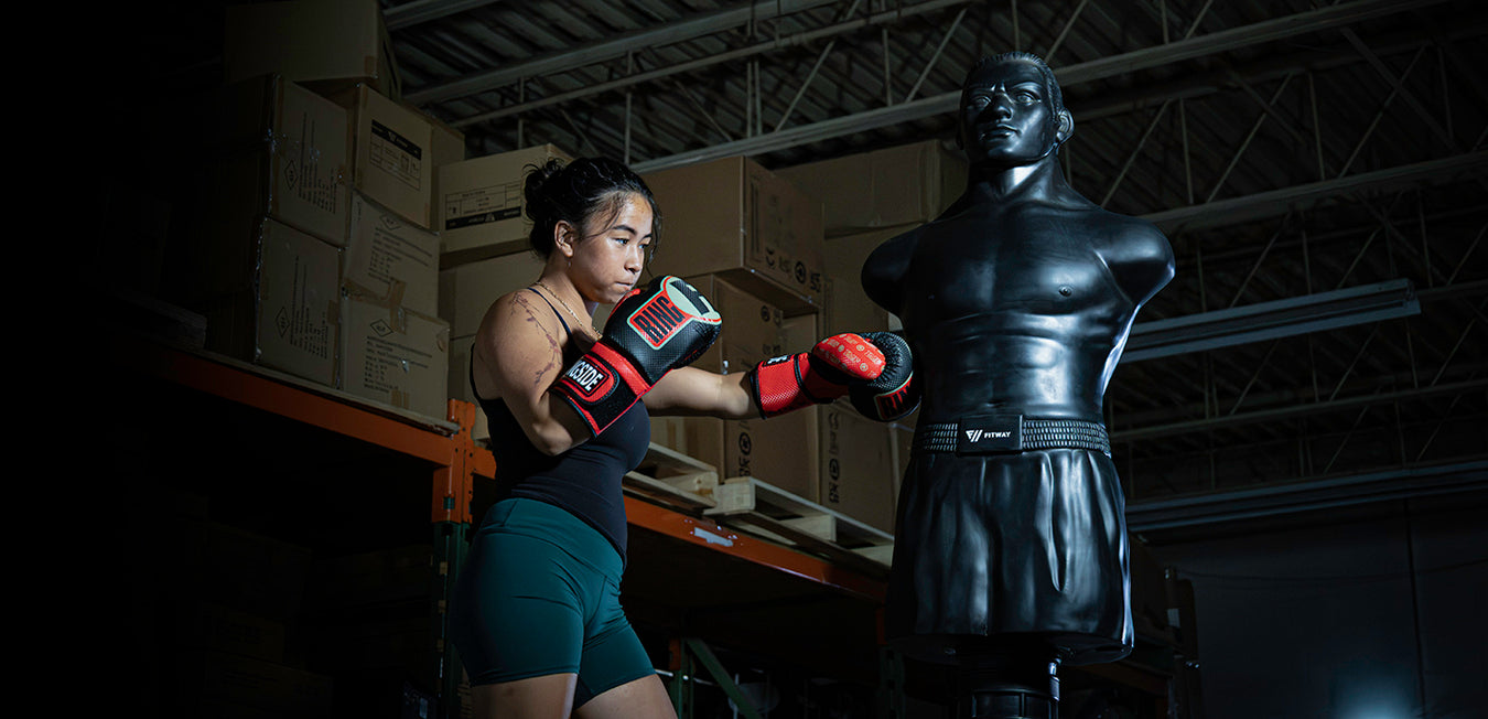 A woman in athletic wear throws a punch at a black boxing dummy in a warehouse setting with cardboard boxes stacked in the background.