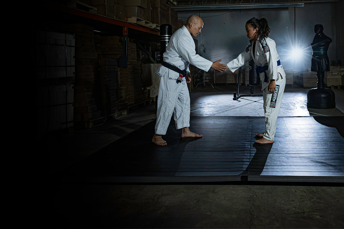 Two martial artists in white Brazilian Jiu-Jitsu uniforms shaking hands on a black mat inside a dimly lit gym or warehouse setting.