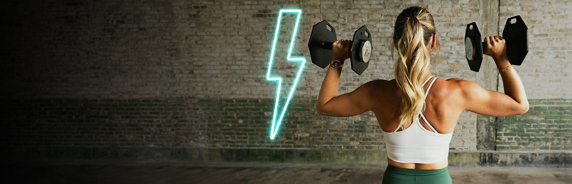 Woman exercising with dumbbells in front of a brick wall with a neon light.