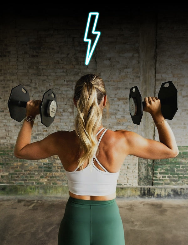 Woman exercising with dumbbells in front of a brick wall with a neon light.