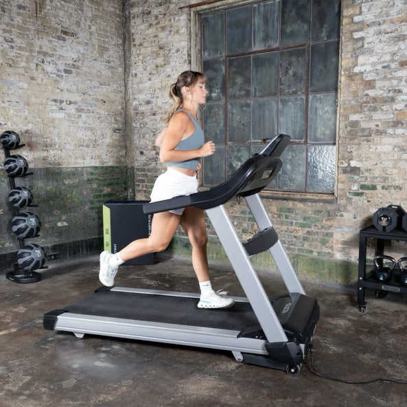 Woman running on a treadmill in an urban setting with brick walls and large windows.